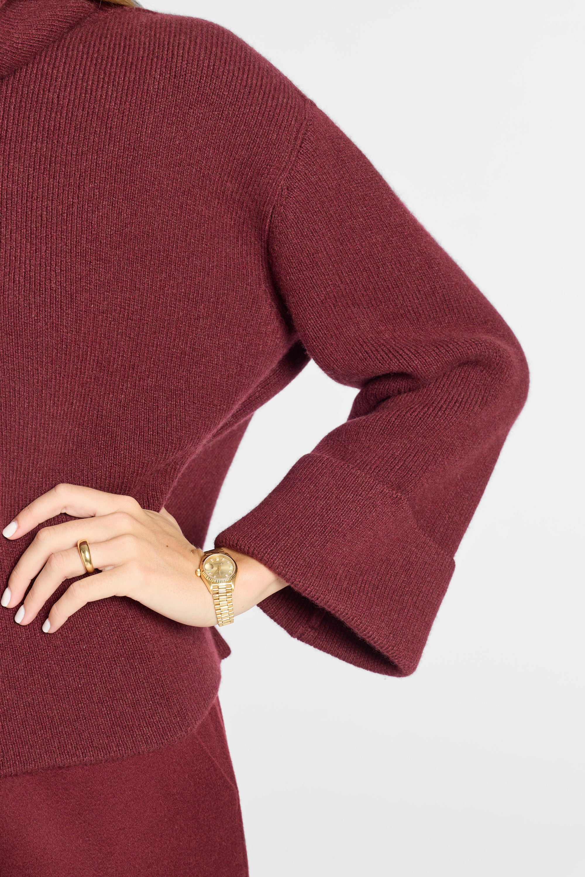A close-up of a person wearing a ribbed knit long-sleeve top or dress in deep burgundy, with one hand resting on the hip. The person is also wearing a gold watch and ring, set against a clean light gray background.