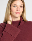 A woman wearing a ribbed knit cowl neck sweater in burgundy, styled with gold hoop earrings and a gold chain bracelet against a light gray background.
