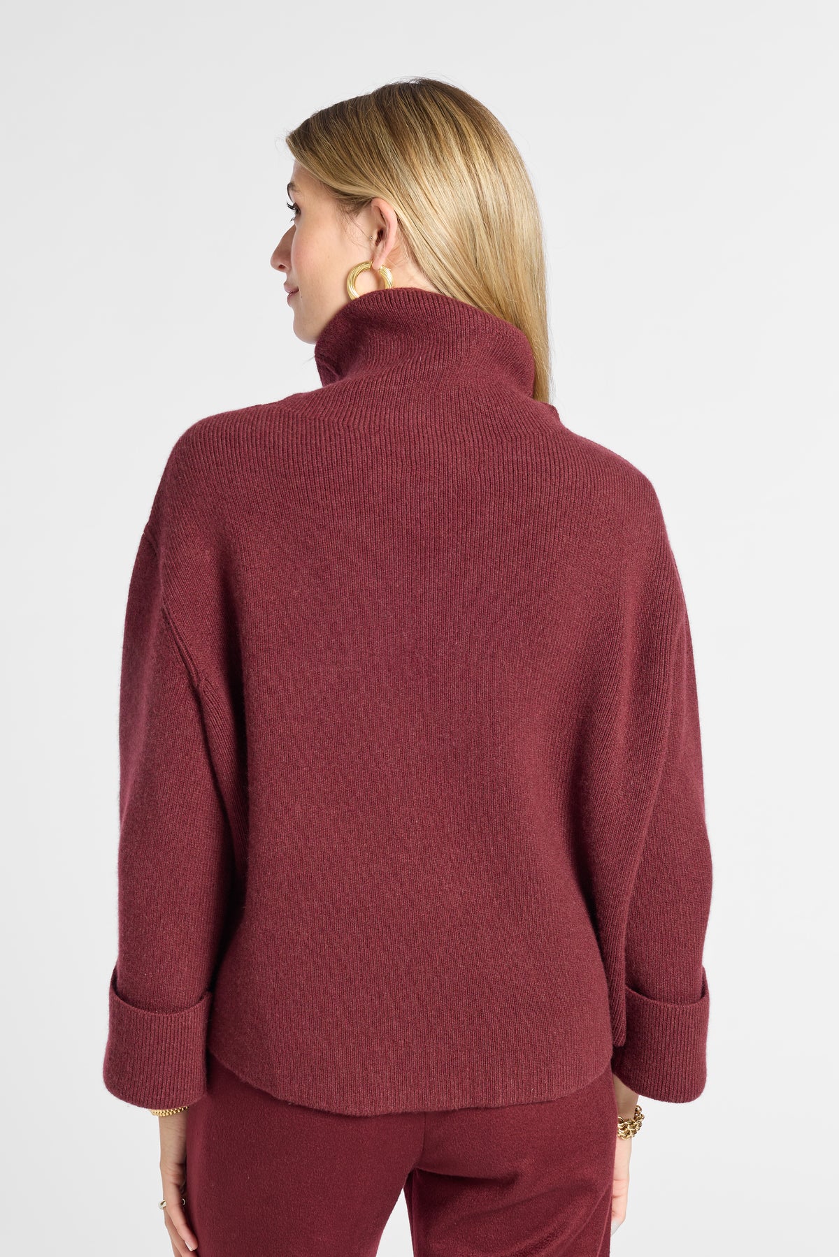 A woman shown from the back wearing a ribbed turtleneck sweater in a deep burgundy color, paired with matching pants, against a plain light gray studio background.