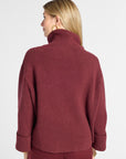 A woman shown from the back wearing a ribbed turtleneck sweater in a deep burgundy color, paired with matching pants, against a plain light gray studio background.