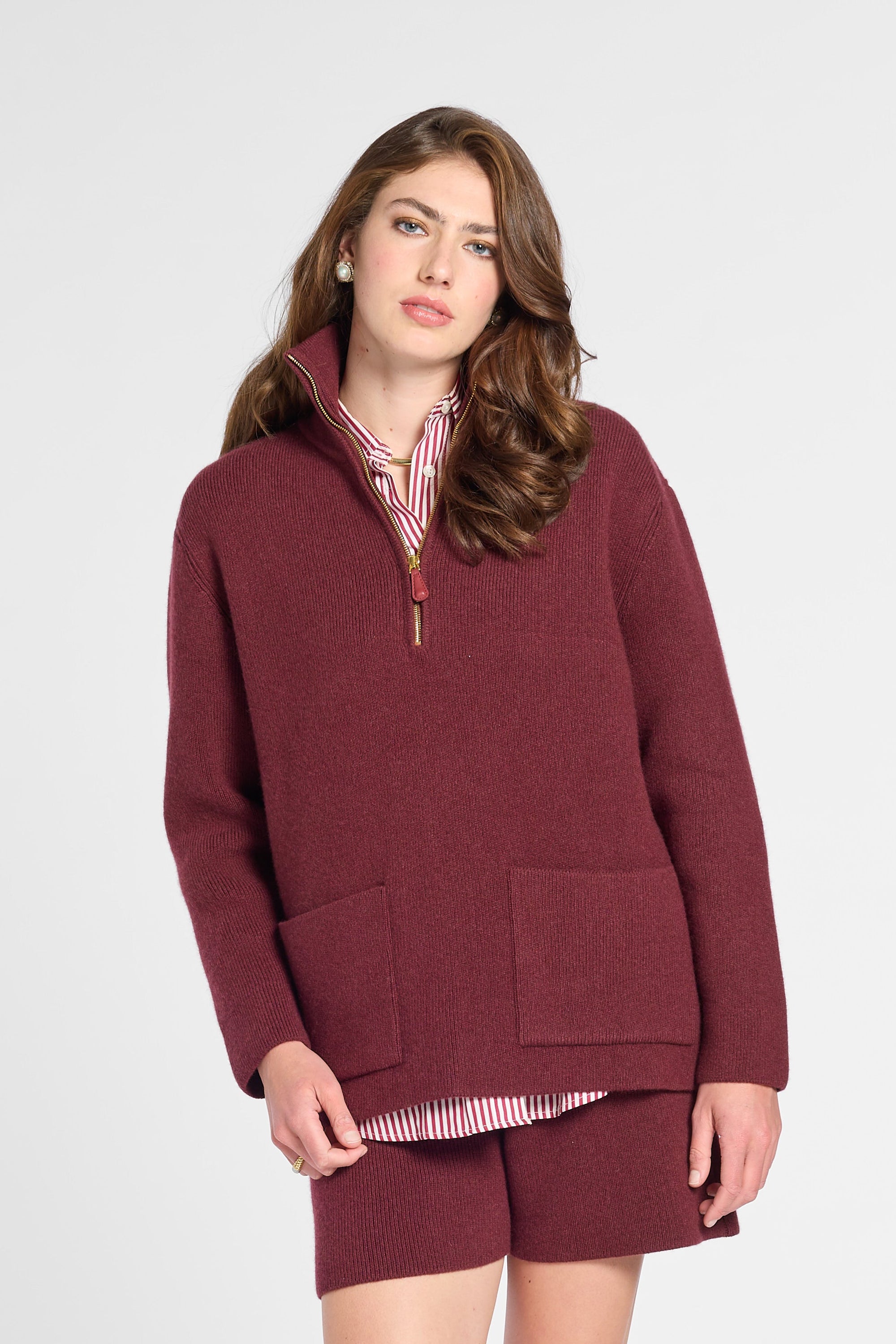 A woman modeling a matching burgundy knit lounge set consisting of a half-zip pullover sweater and shorts, styled over a pink striped shirt and brown leather shoes, against a light gray studio background.