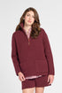 A woman modeling a matching burgundy knit lounge set consisting of a half-zip pullover sweater and shorts, styled over a pink striped shirt and brown leather shoes, against a light gray studio background.