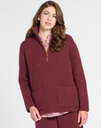 A woman modeling a matching burgundy knit lounge set consisting of a half-zip pullover sweater and shorts, styled over a pink striped shirt and brown leather shoes, against a light gray studio background.