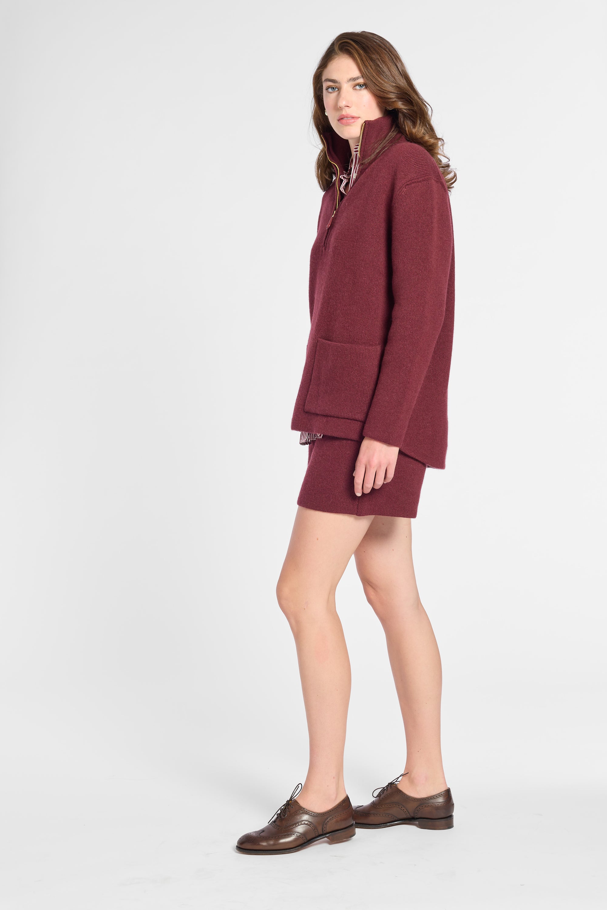 A woman modeling a matching burgundy knit set consisting of a mock-neck zip-up jacket and a short skirt, paired with brown leather oxford shoes, against a light gray studio background.
