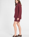 A woman modeling a matching burgundy knit set consisting of a mock-neck zip-up jacket and a short skirt, paired with brown leather oxford shoes, against a light gray studio background.