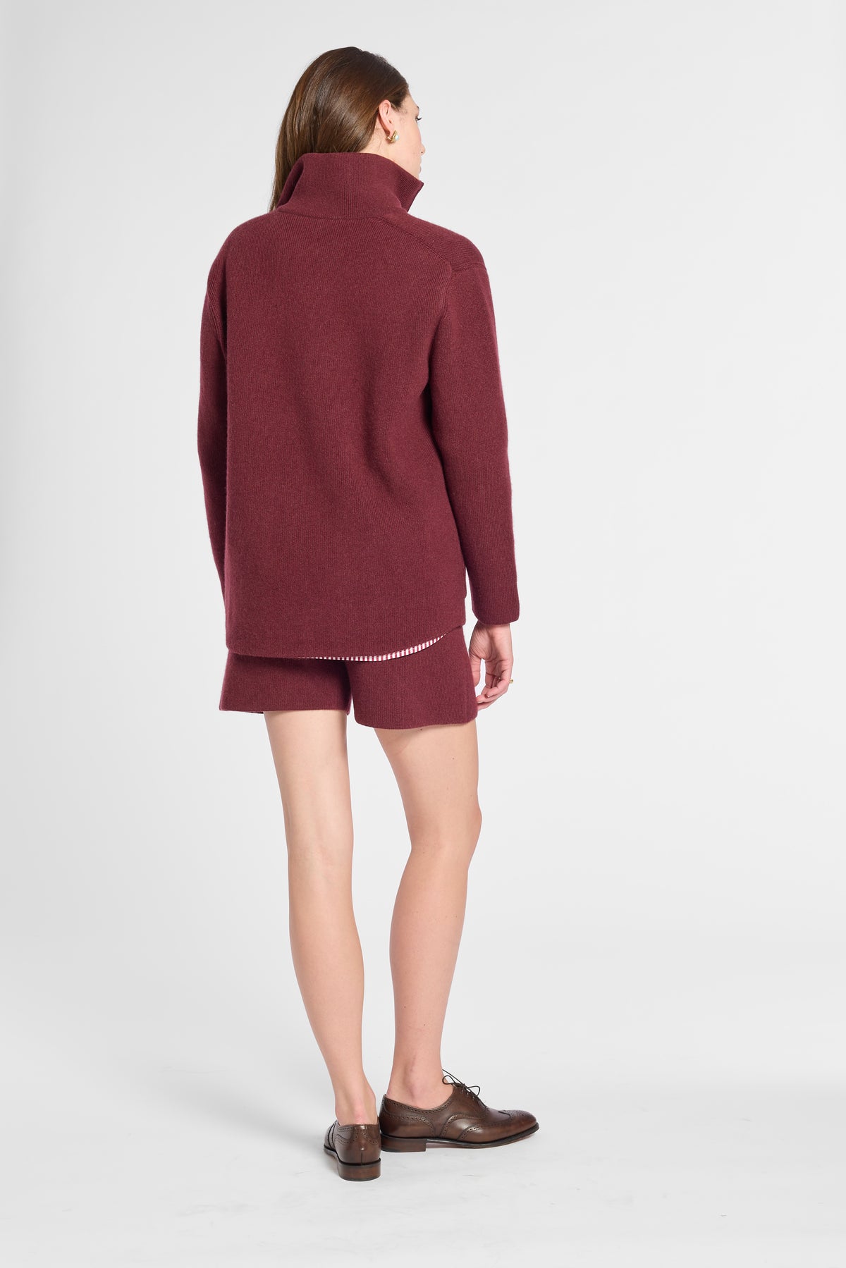A woman shown from the back wearing a matching burgundy knit turtleneck sweater and shorts set, paired with brown leather shoes, against a light gray studio background.