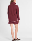 A woman shown from the back wearing a matching burgundy knit turtleneck sweater and shorts set, paired with brown leather shoes, against a light gray studio background.