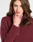 A woman wearing a ribbed maroon half-zip sweater over a striped shirt, photographed against a plain light gray studio background.