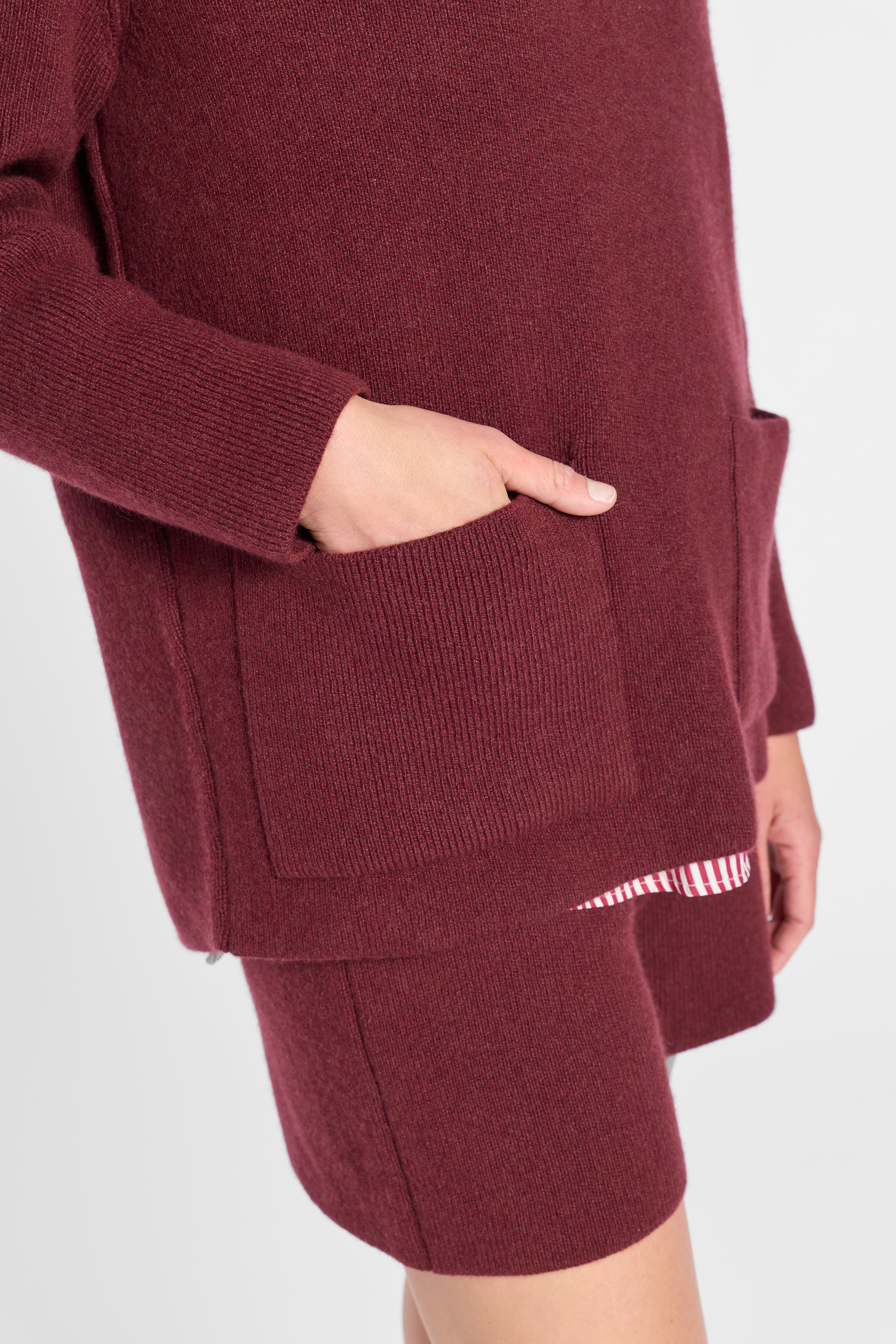 A close-up of a person wearing a ribbed knit quarter zip sweater and matching shorts in a deep burgundy color, with a hand tucked into a pocket and a striped hem detail visible.