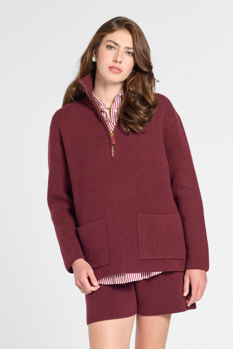 A woman modeling a matching burgundy knit lounge set consisting of a half-zip pullover sweater and shorts, styled over a pink striped shirt and brown leather shoes, against a light gray studio background.