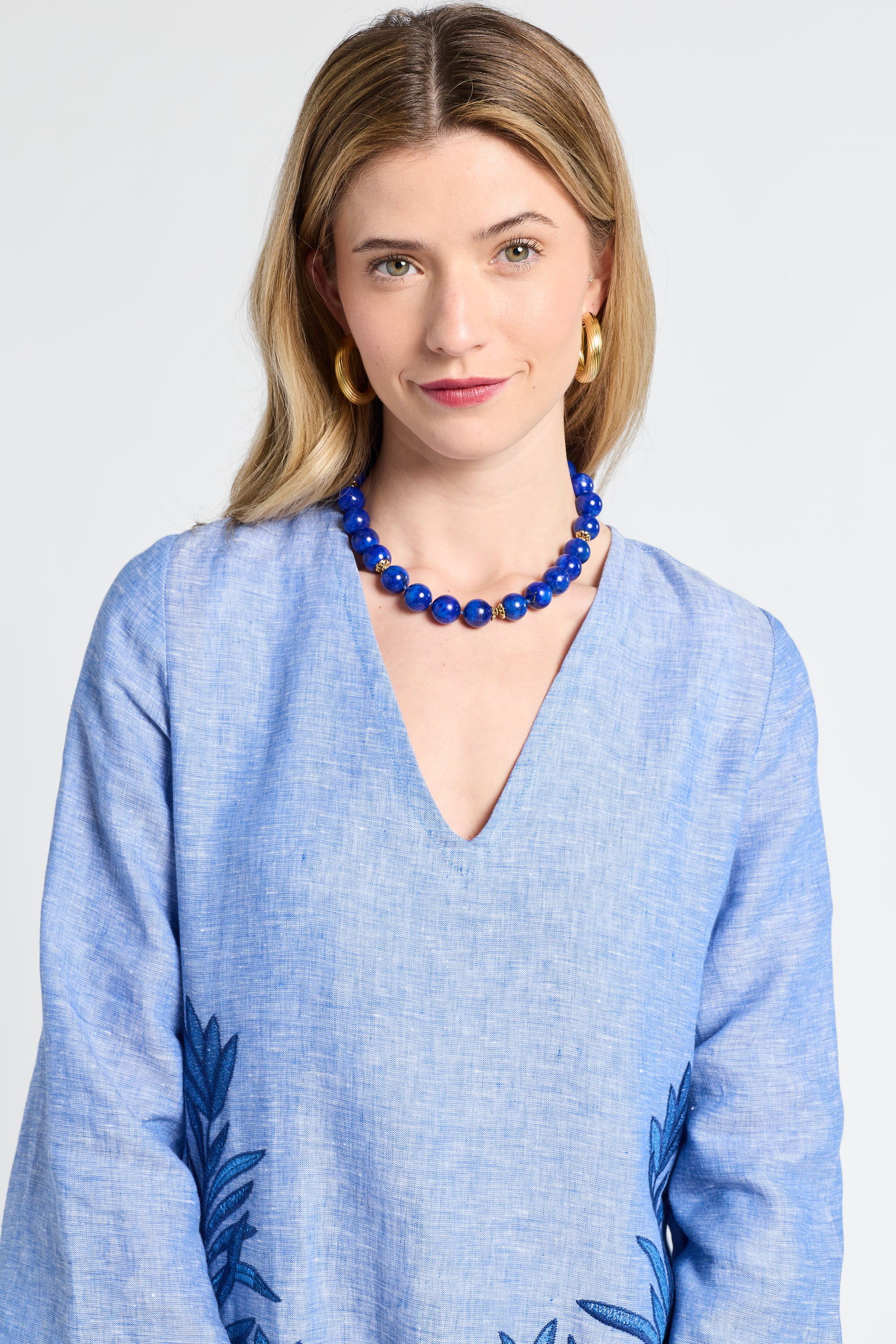Woman wearing a blue caftan with floral patterns and a blue necklace against a white background