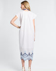 Woman wearing a white dress with blue embroidery details on a plain background