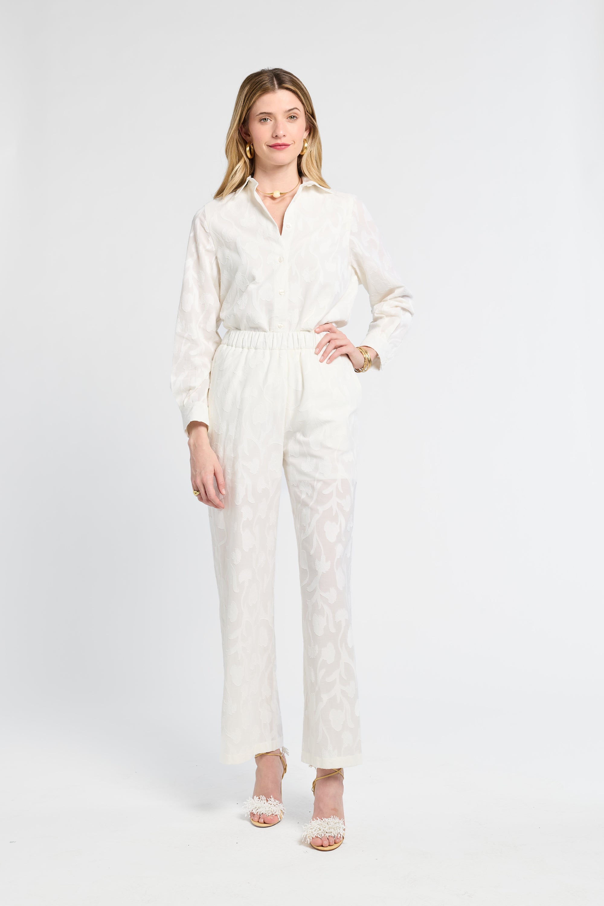 Woman modeling white pants with floral detailing against a white backdrop