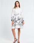 Woman modeling a long sleeve white dress with black floral detailing along the bottom on a white backdrop