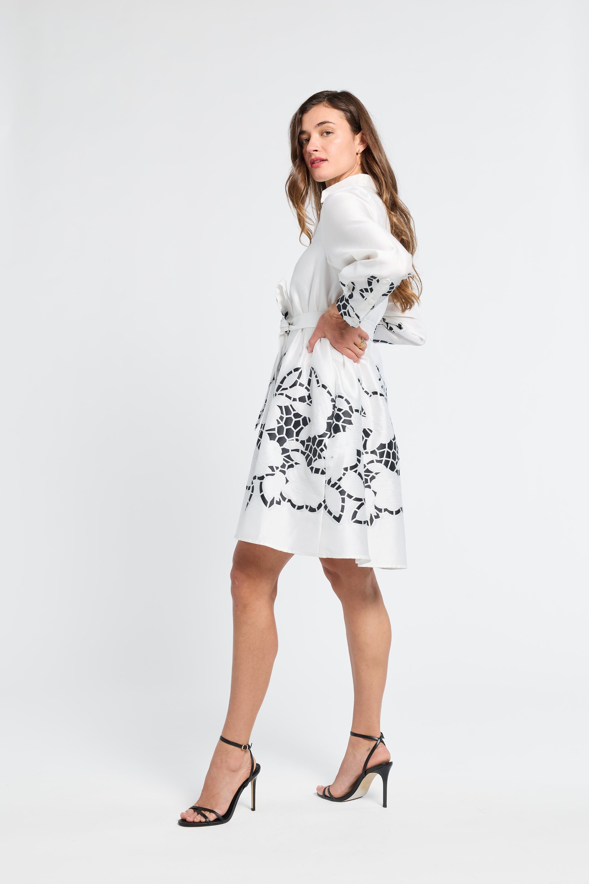 Lady wearing a long sleeve white dress with black floral detailing and black heels on a white backdrop