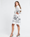 Lady wearing a long sleeve white dress with black floral detailing and black heels on a white backdrop