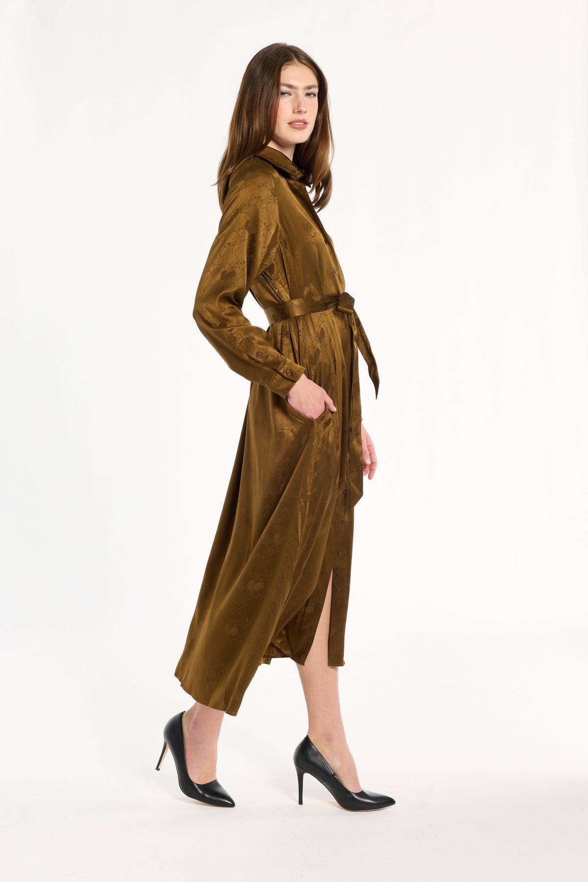 A woman modeling the Golden Umber Eleanor Shirt Dress with a tied sash and black heels, side view, photographed against a plain white studio background.