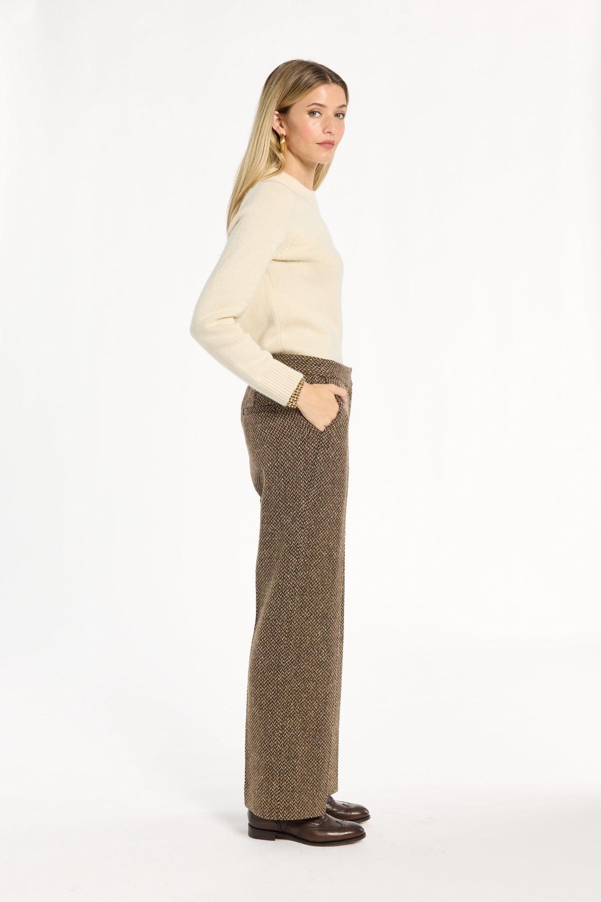 A woman modeling the Tobacco Tweed Ellis Pants with a cream sweater and brown loafers, side view, photographed against a plain white studio background.