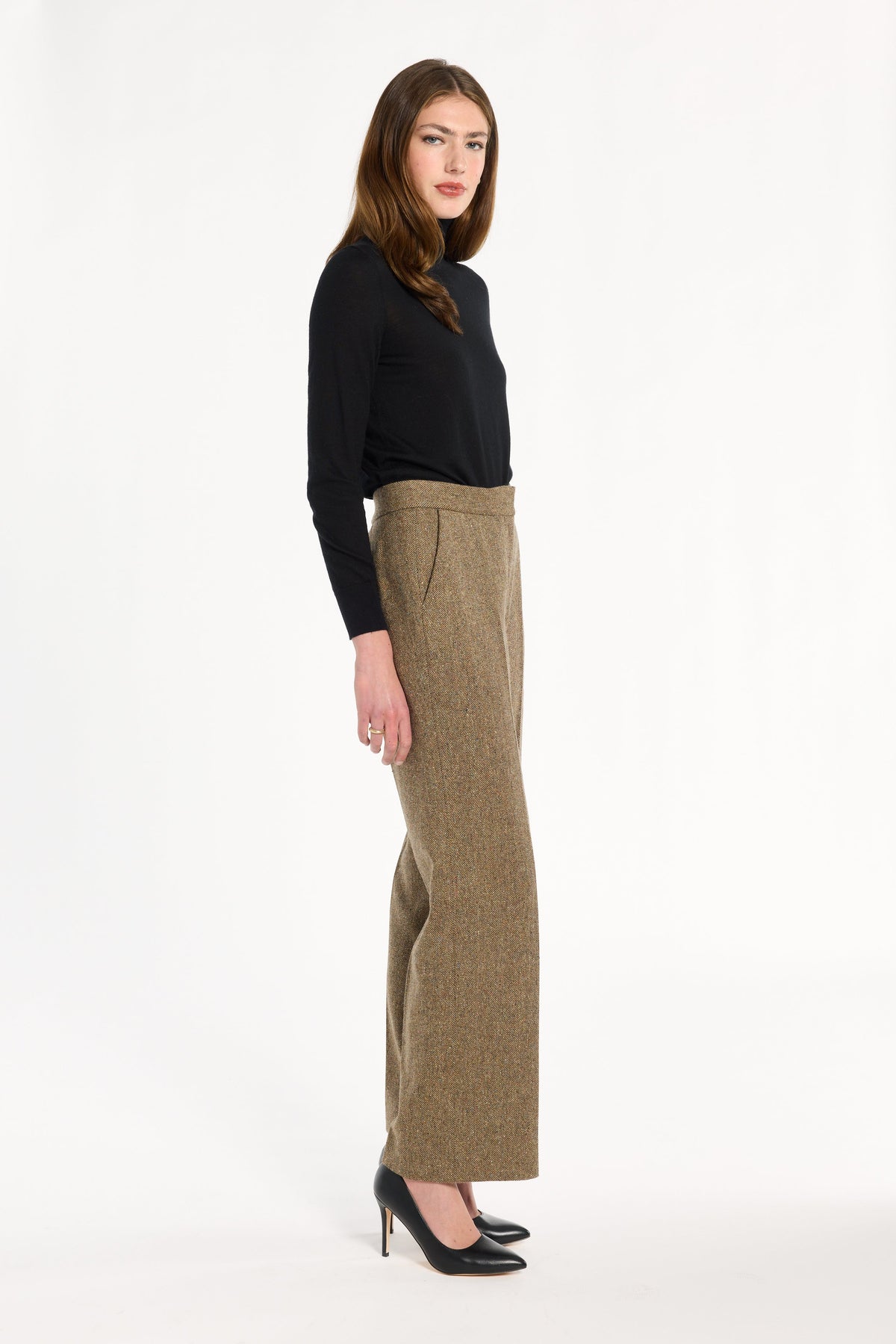 A woman modeling the Painter's Fleck Herringbone Ellis Pants with a black turtleneck and heels, side view, photographed against a plain white studio background.