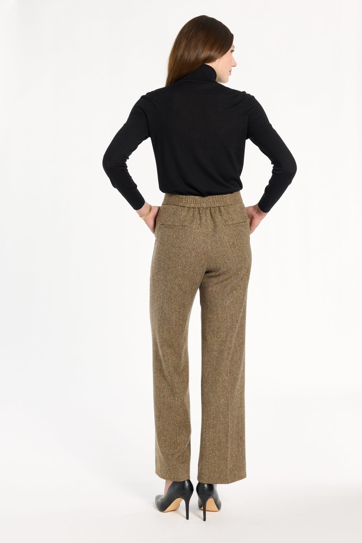 A woman modeling the Painter's Fleck Herringbone Ellis Pants with a black turtleneck and heels, back view, photographed against a plain white studio background.