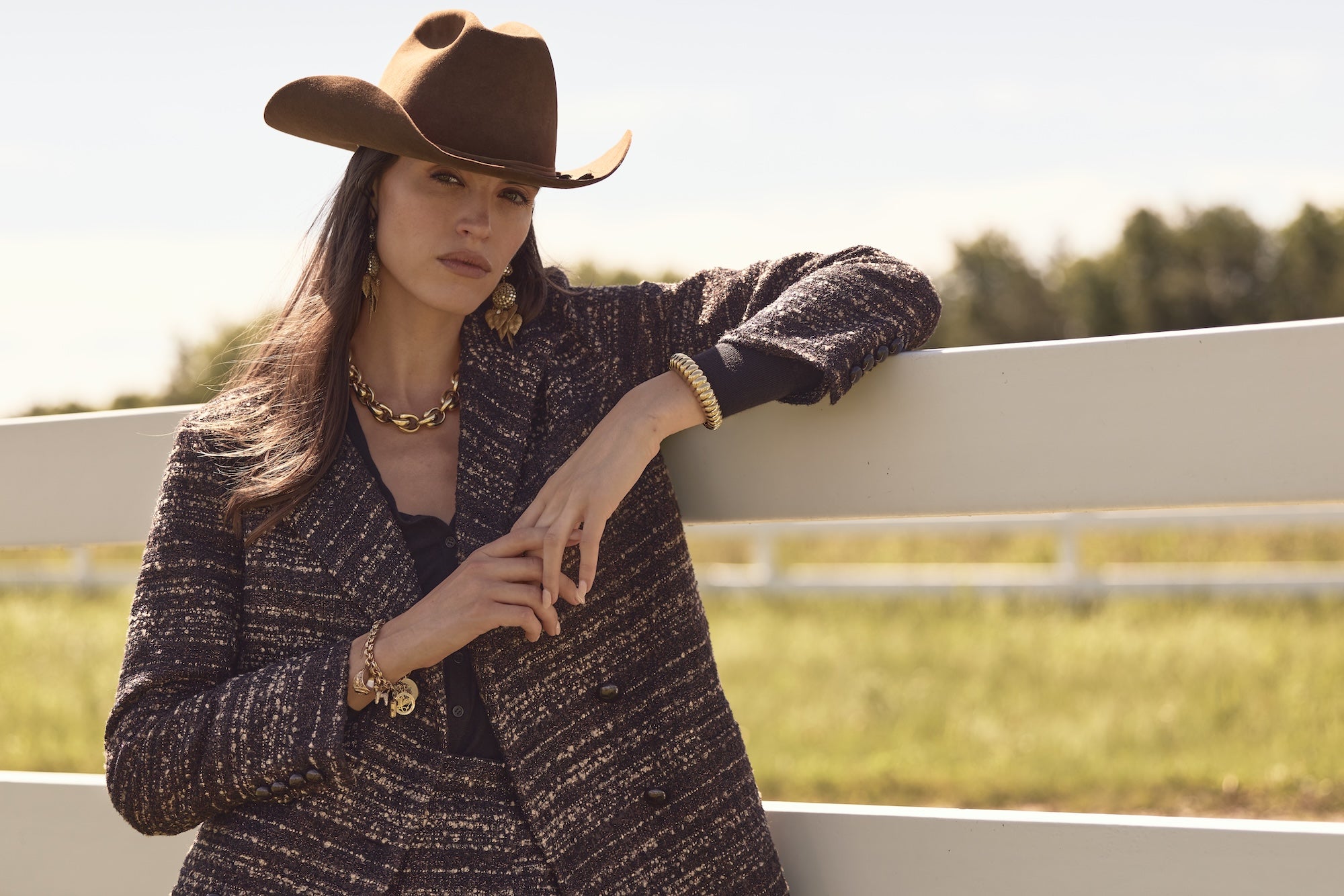 Woman wearing a brown cowboy hat and coat, standing by a fence with a field and trees in the background.