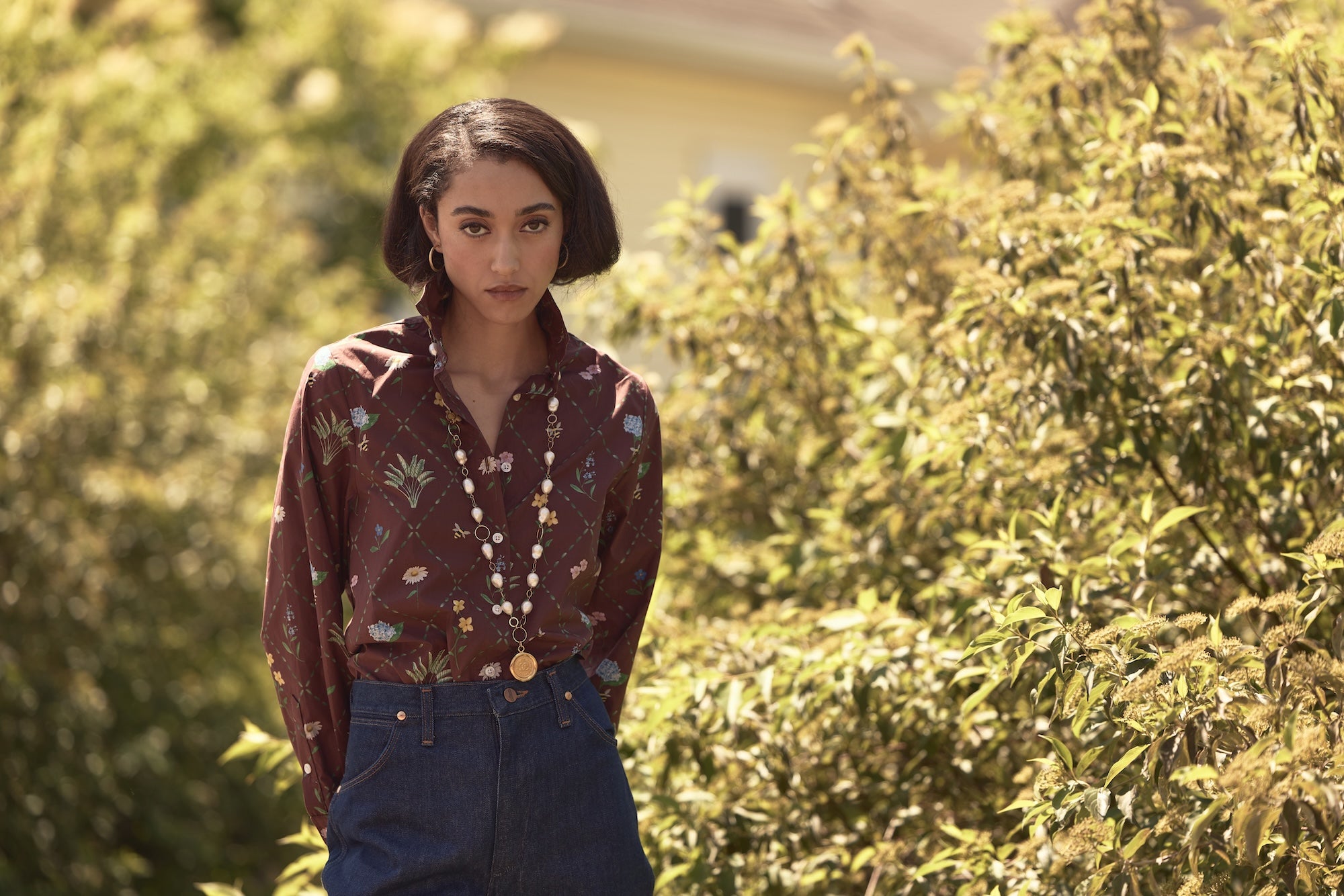 Woman wearing a patterned shirt and blue jeans standing in a garden.