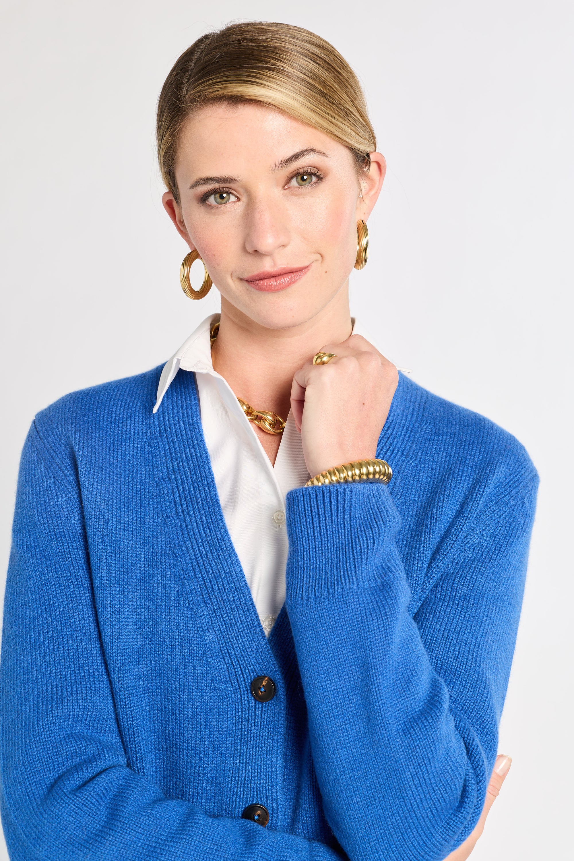Woman wearing a blue cardigan over a white shirt with gold jewelry on a light gray background