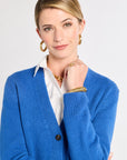 Woman wearing a blue cardigan over a white shirt with gold jewelry on a light gray background