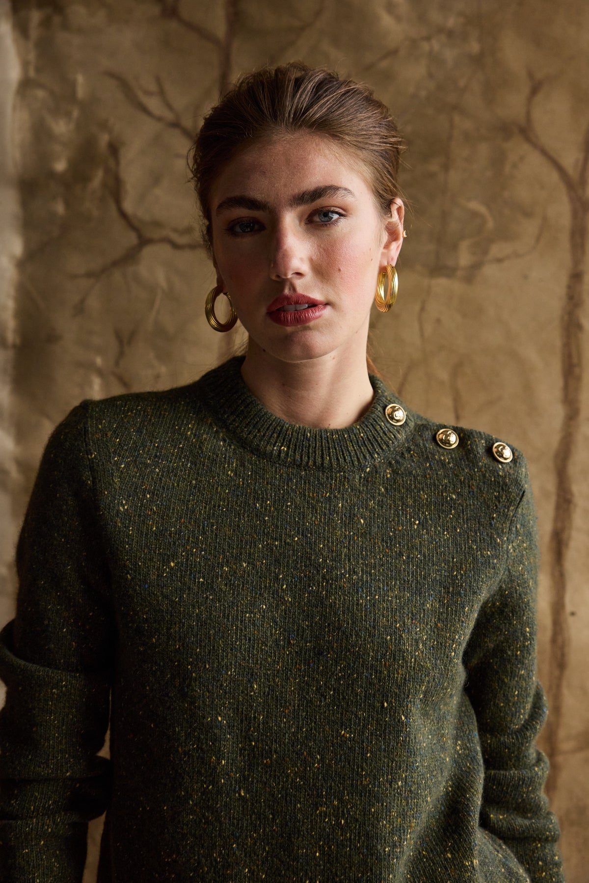 Woman modeling a flecked green cashmere crewneck with gold buttons and a tan background