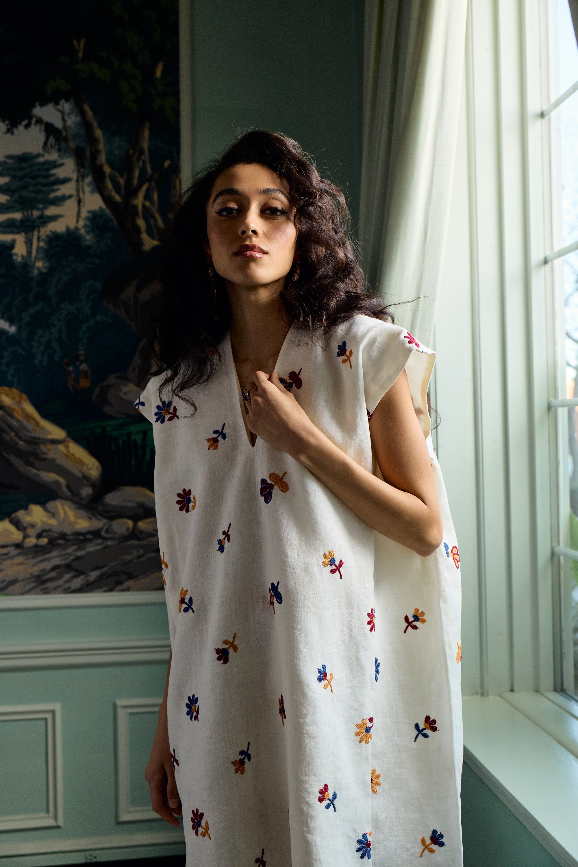 Woman wearing a white sleeveless dress with colorful floral patterns in a room with a painting and window.
