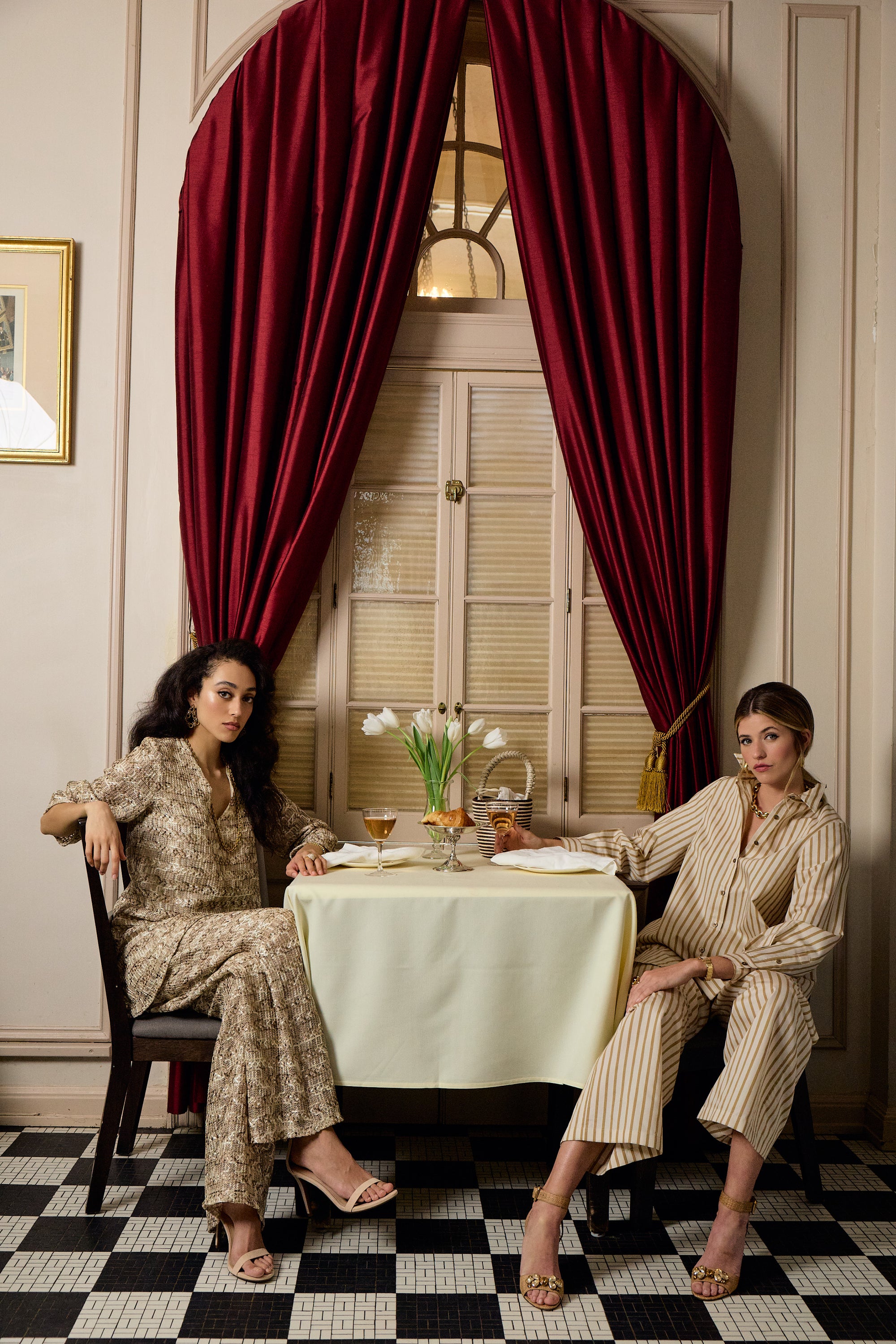 Two women sitting at a table in a room with red curtains and checkered floor.