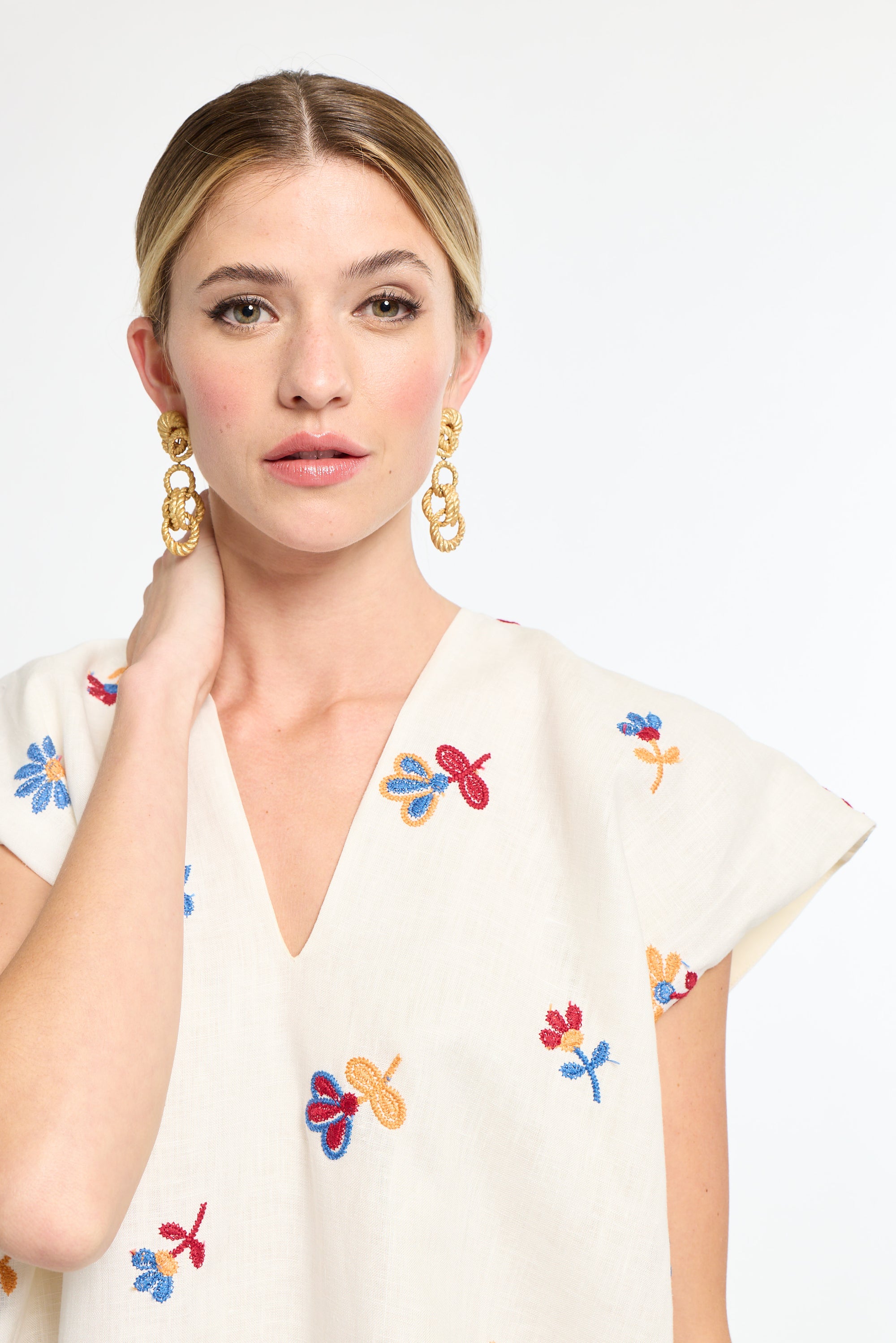 Woman wearing a white top with colorful floral embroidery and gold earrings on a white background