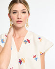 Woman wearing a white top with colorful floral embroidery and gold earrings on a white background