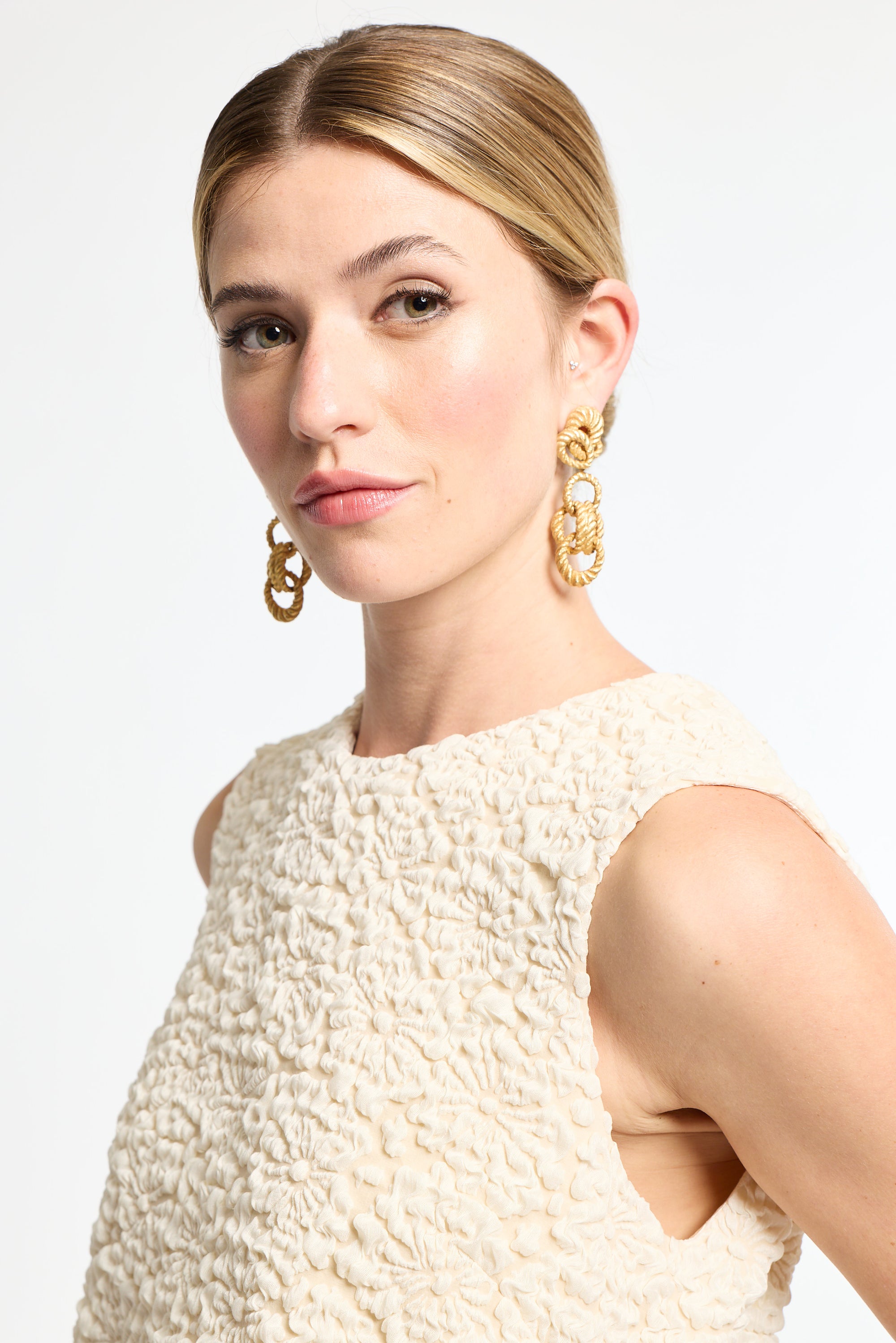 Woman wearing gold earrings and a textured cream dress against a white background