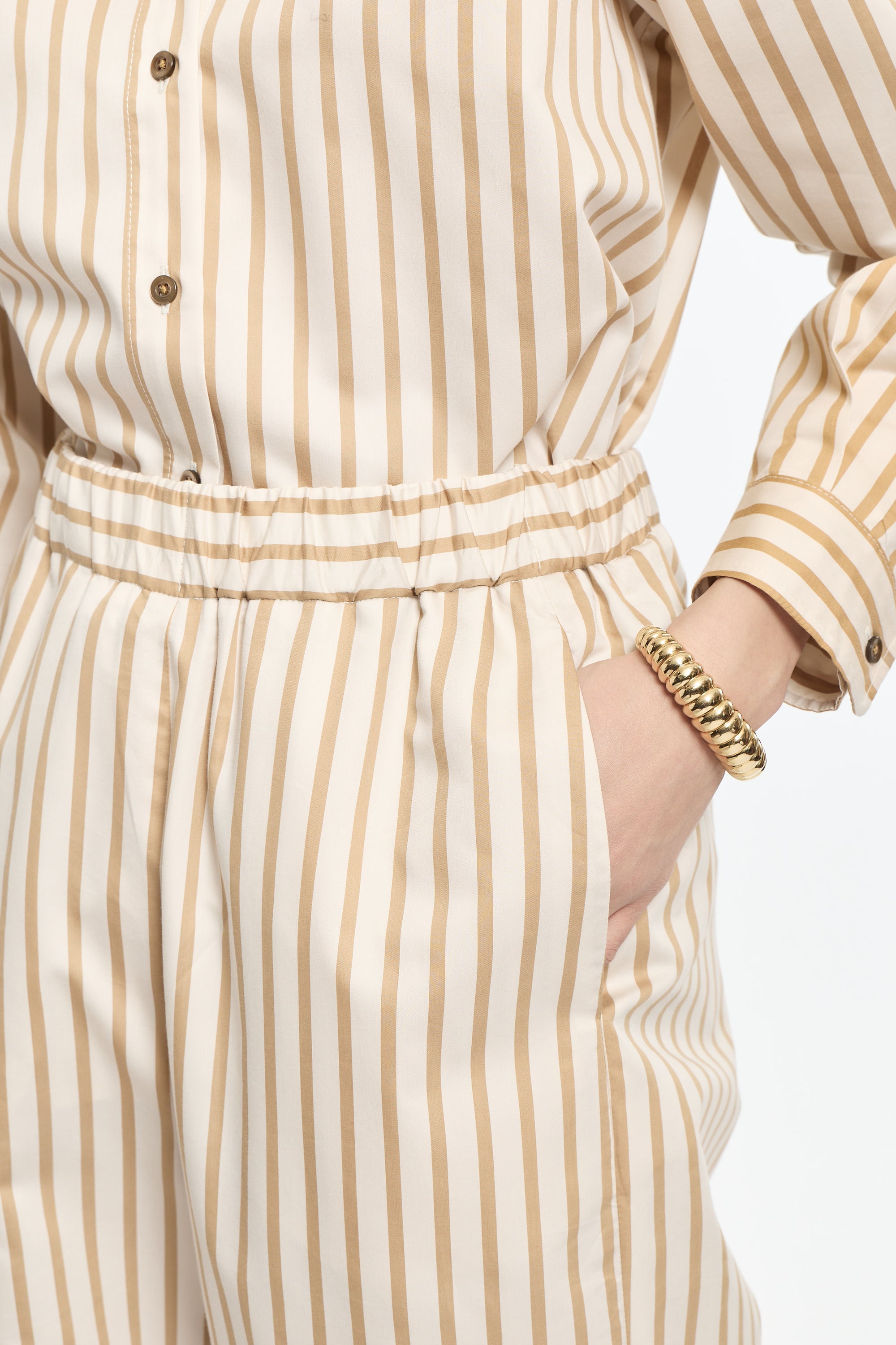 Person wearing a beige and white striped outfit with a gold bracelet on a plain background