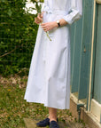 A person wearing a white and light blue vertically striped midi dress with a tie waist, standing outdoors next to a teal wooden shed and holding white flowers.