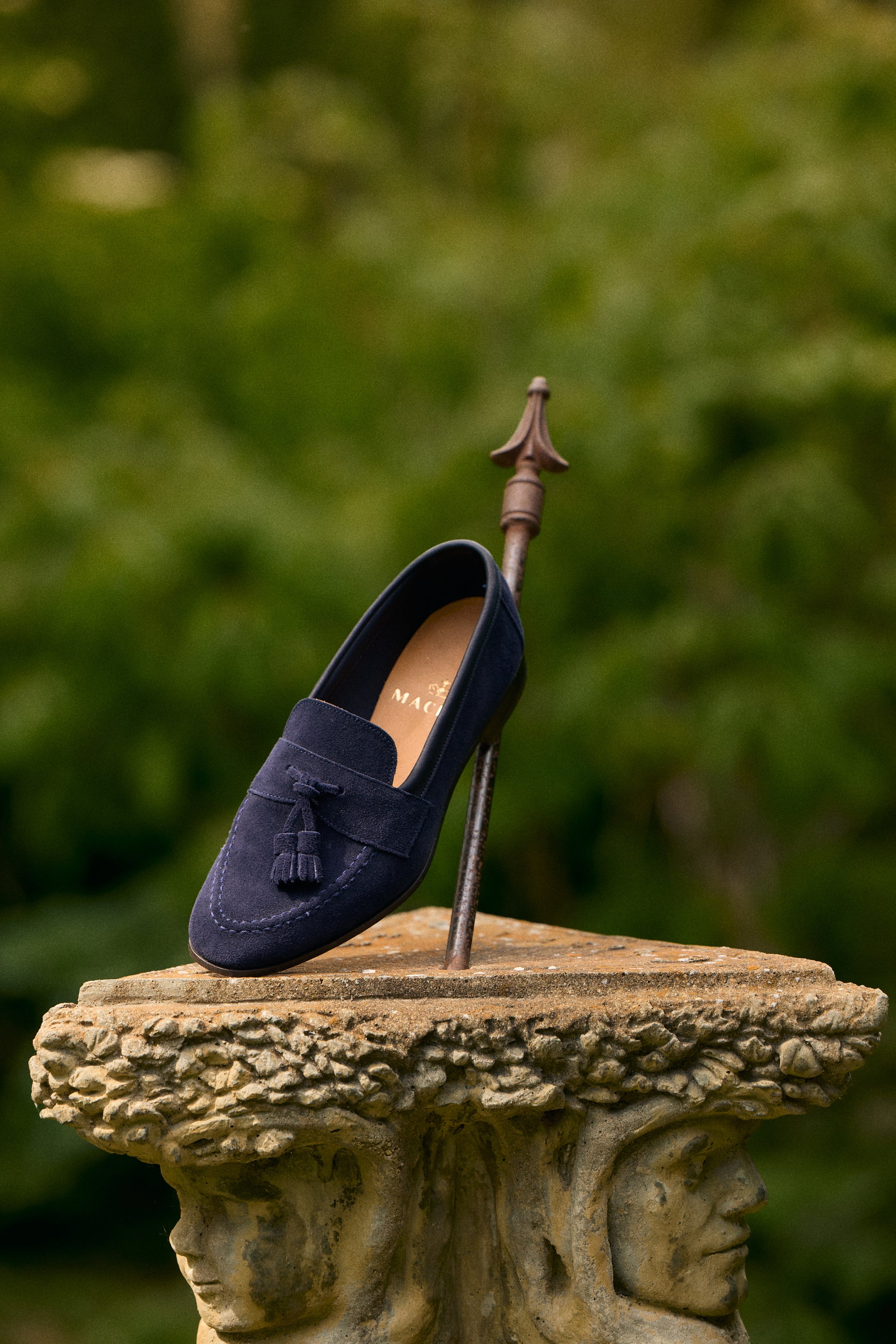 A navy blue suede tassel loafer shoe displayed on a metal stand atop a stone pedestal, with a soft-focus green garden background.