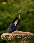 A navy blue suede tassel loafer shoe displayed on a metal stand atop a stone pedestal, with a soft-focus green garden background.