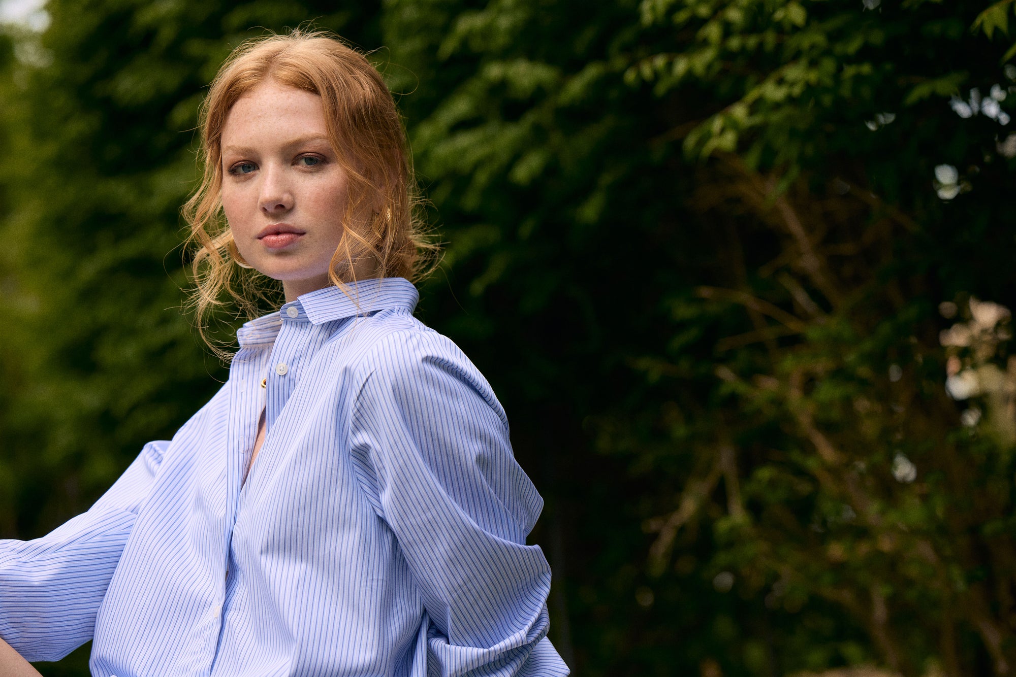Woman wearing a light blue striped shirt outdoors with greenery in the background