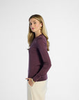 side profile of a woman wearing a purple long-sleeve top and beige pants on a white background
