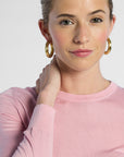 close up of a woman wearing gold hoop earrings and a pink sweater against a white background