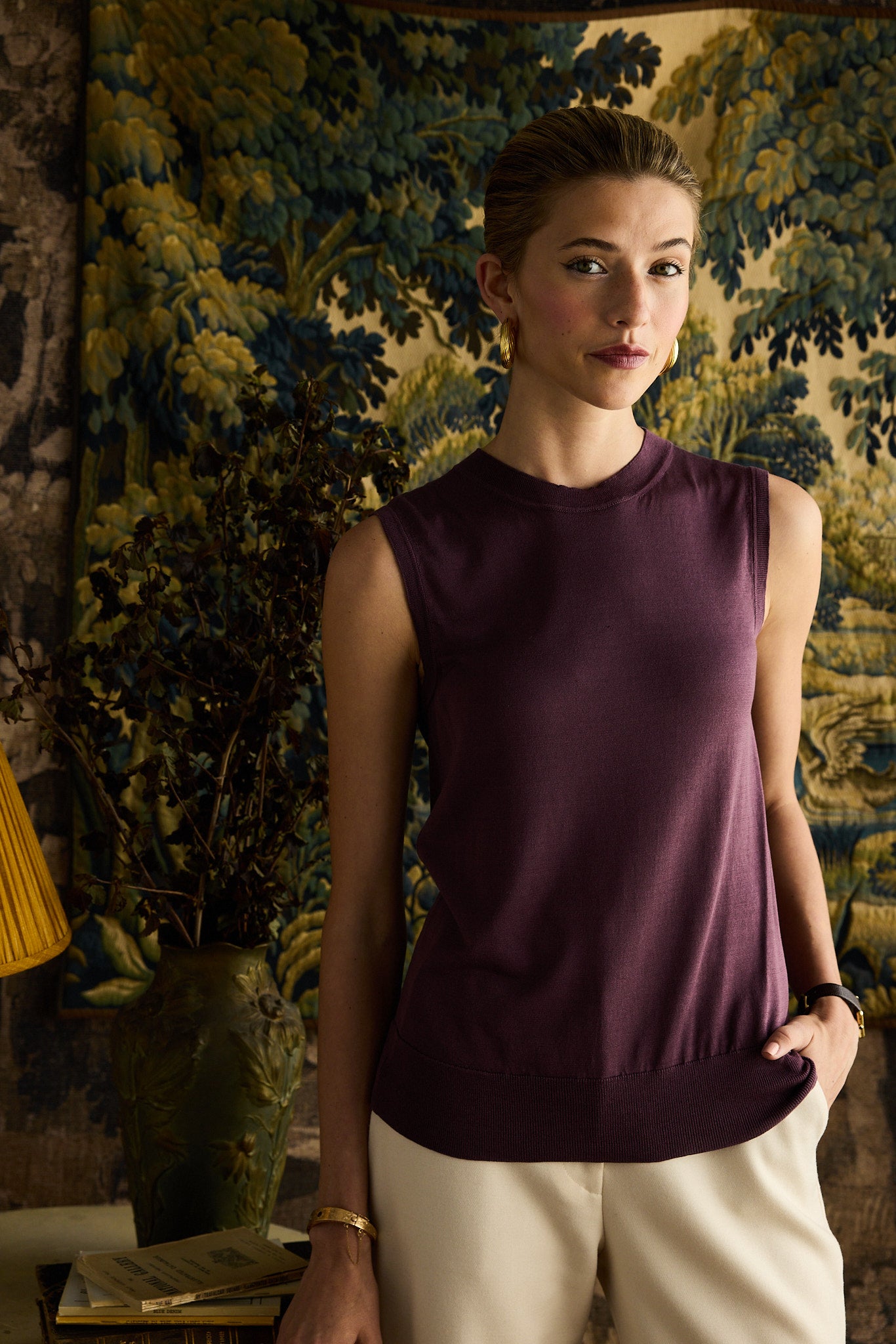 Woman wearing a purple sleeveless top and beige pants standing in front of a decorative wall.
