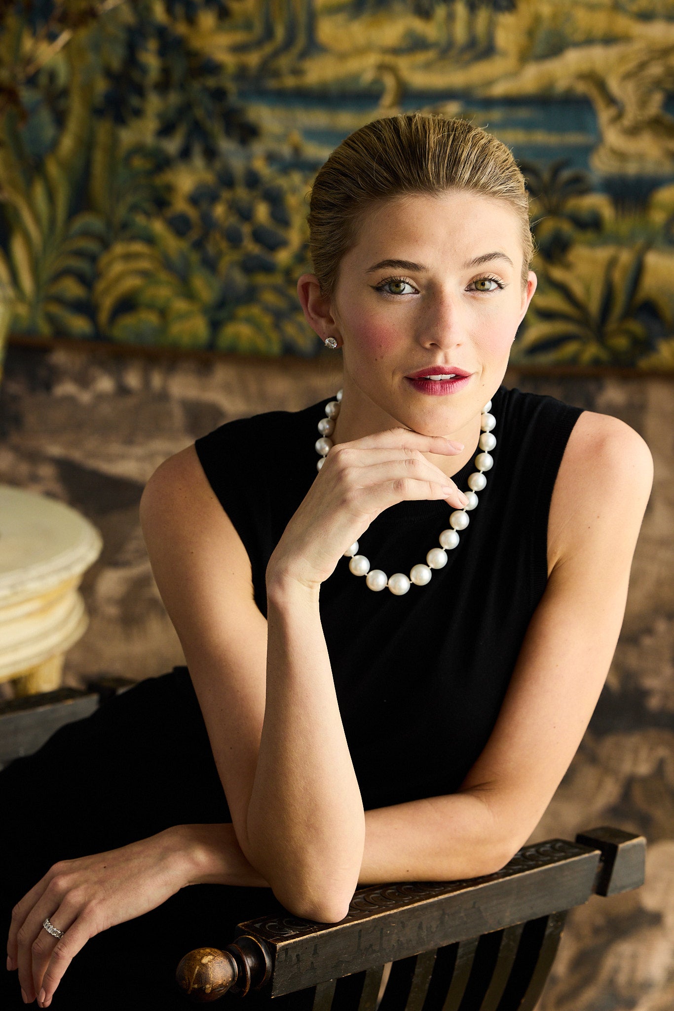 Woman in a black silk shell with a pearl necklace against a decorative wall.