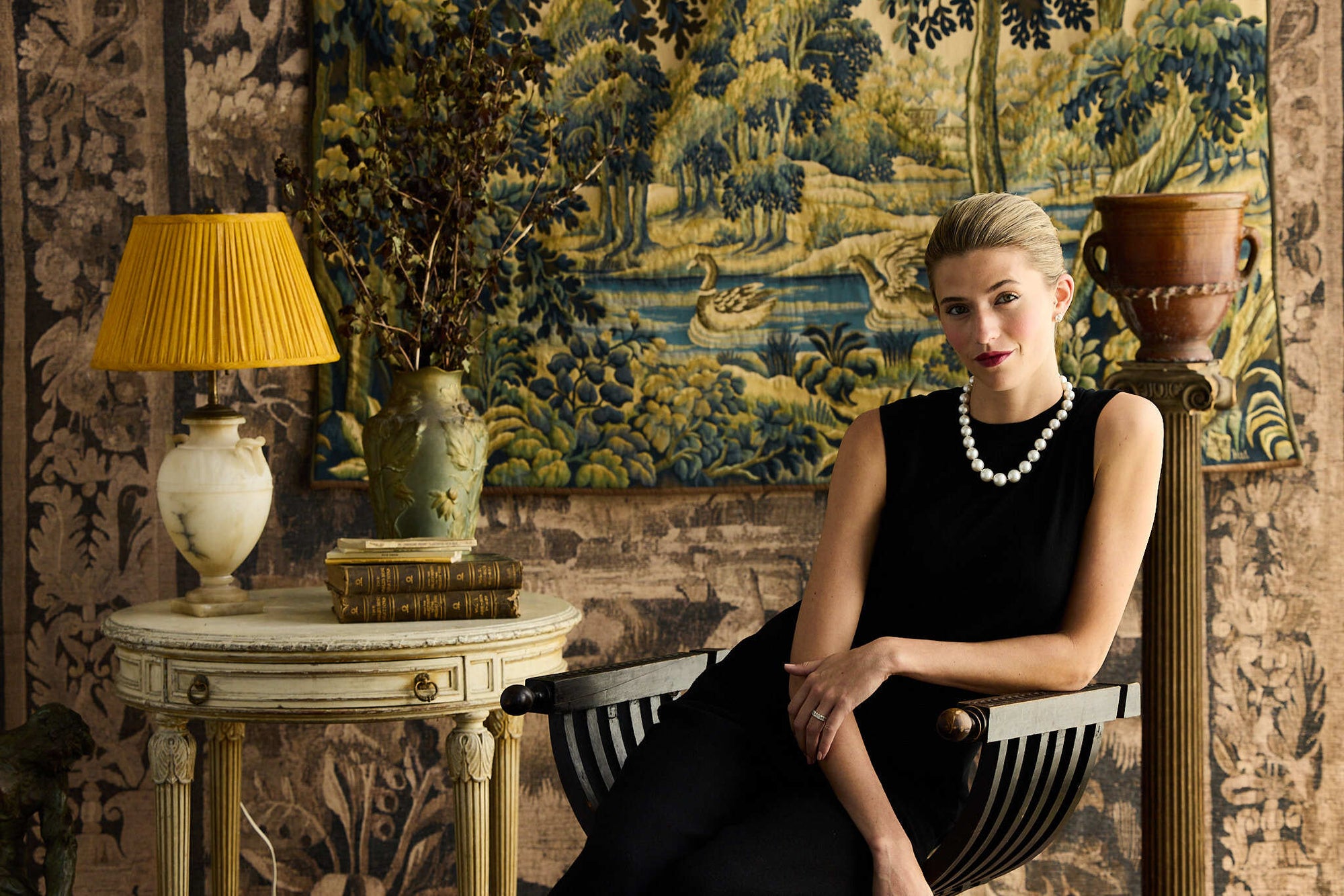 Woman in a black silk knit shell sitting in an ornate room with decorative wall tapestry and lamp.