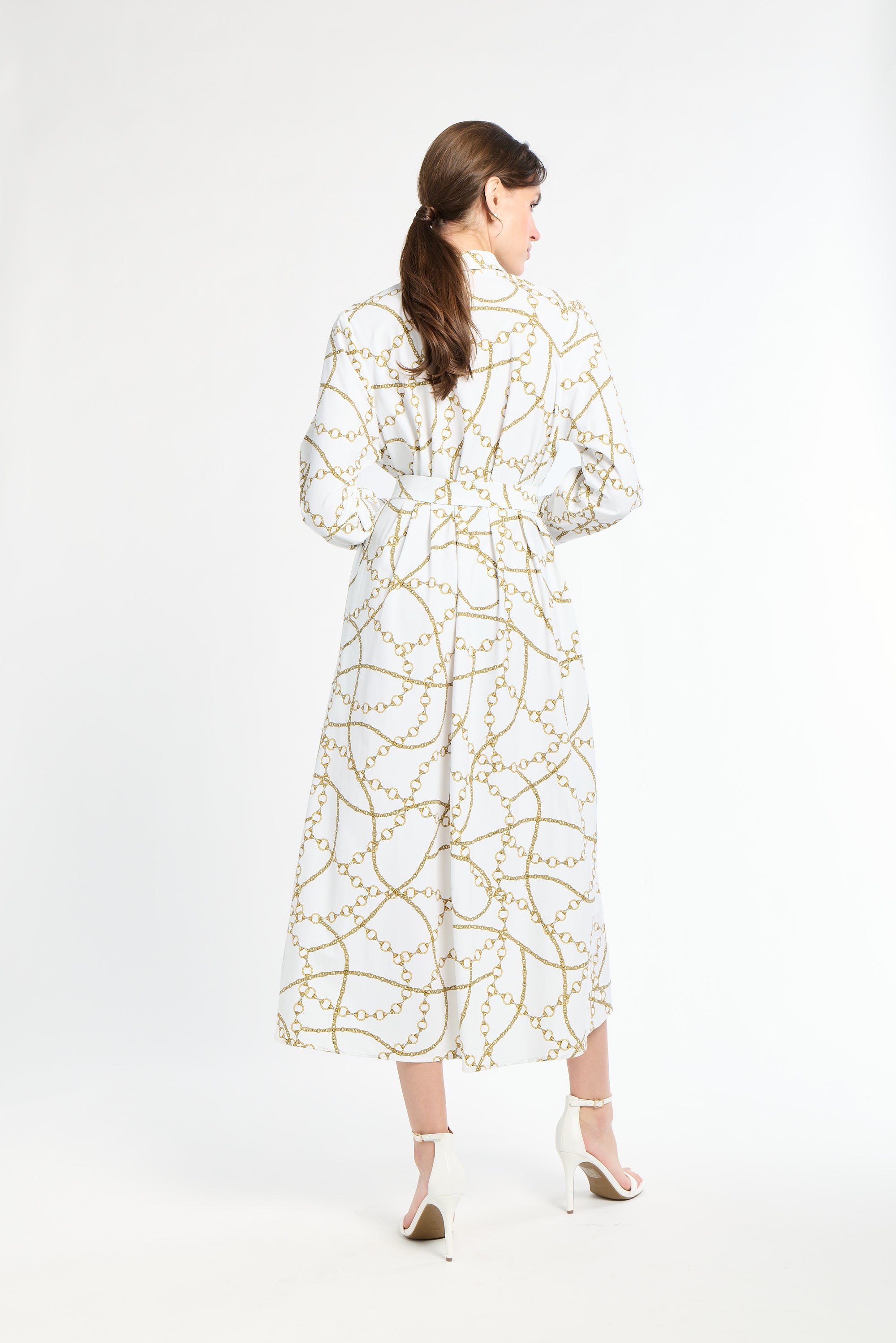 Woman wearing a long sleeve white dress with a gold chain pattern on a white back drop
