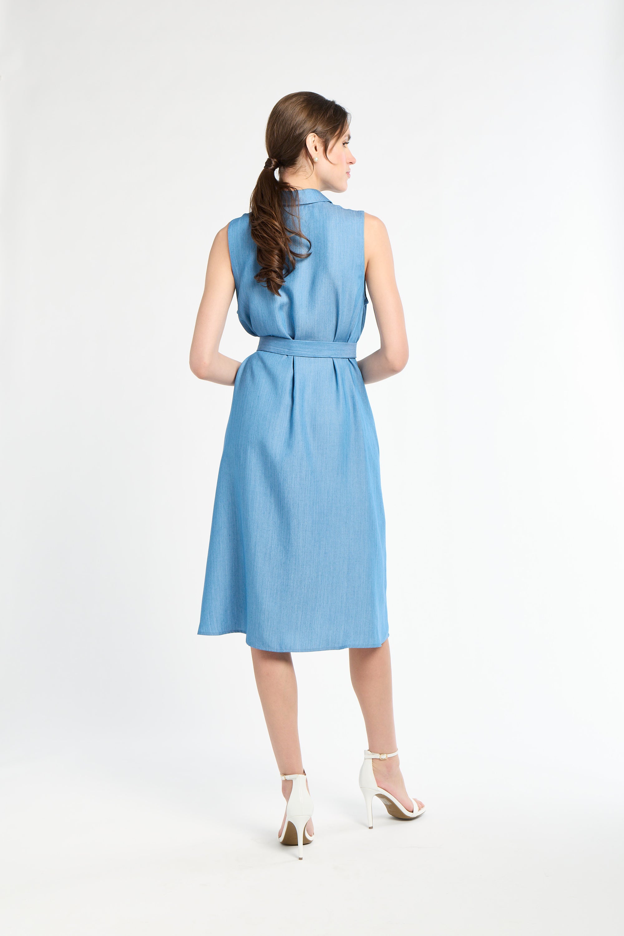 Woman with dark hair modeling blue dress  and wearing white heels against white backdrop