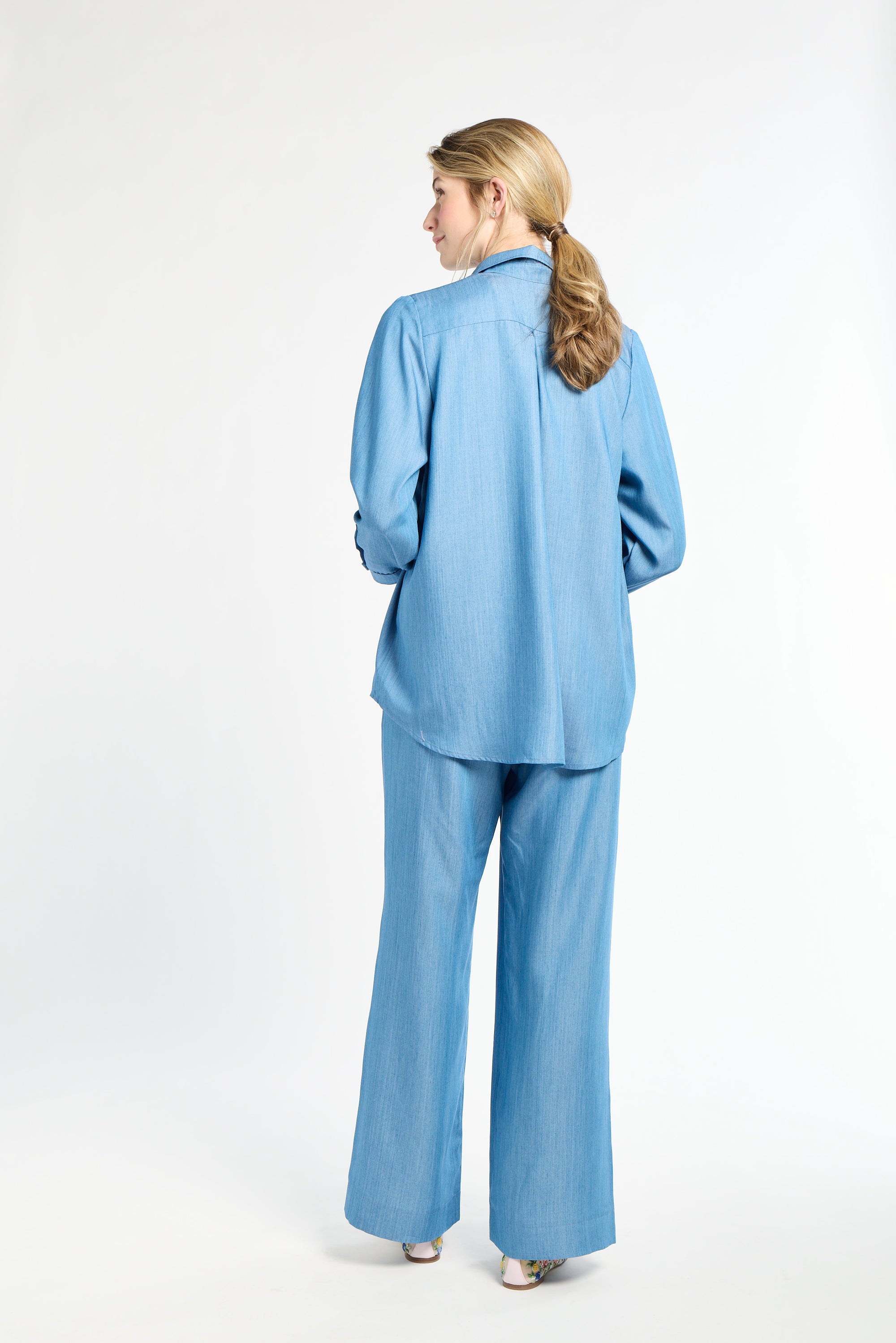 Woman standing against a white backdrop wearing a long sleeve blue shirt and long sleeve pants