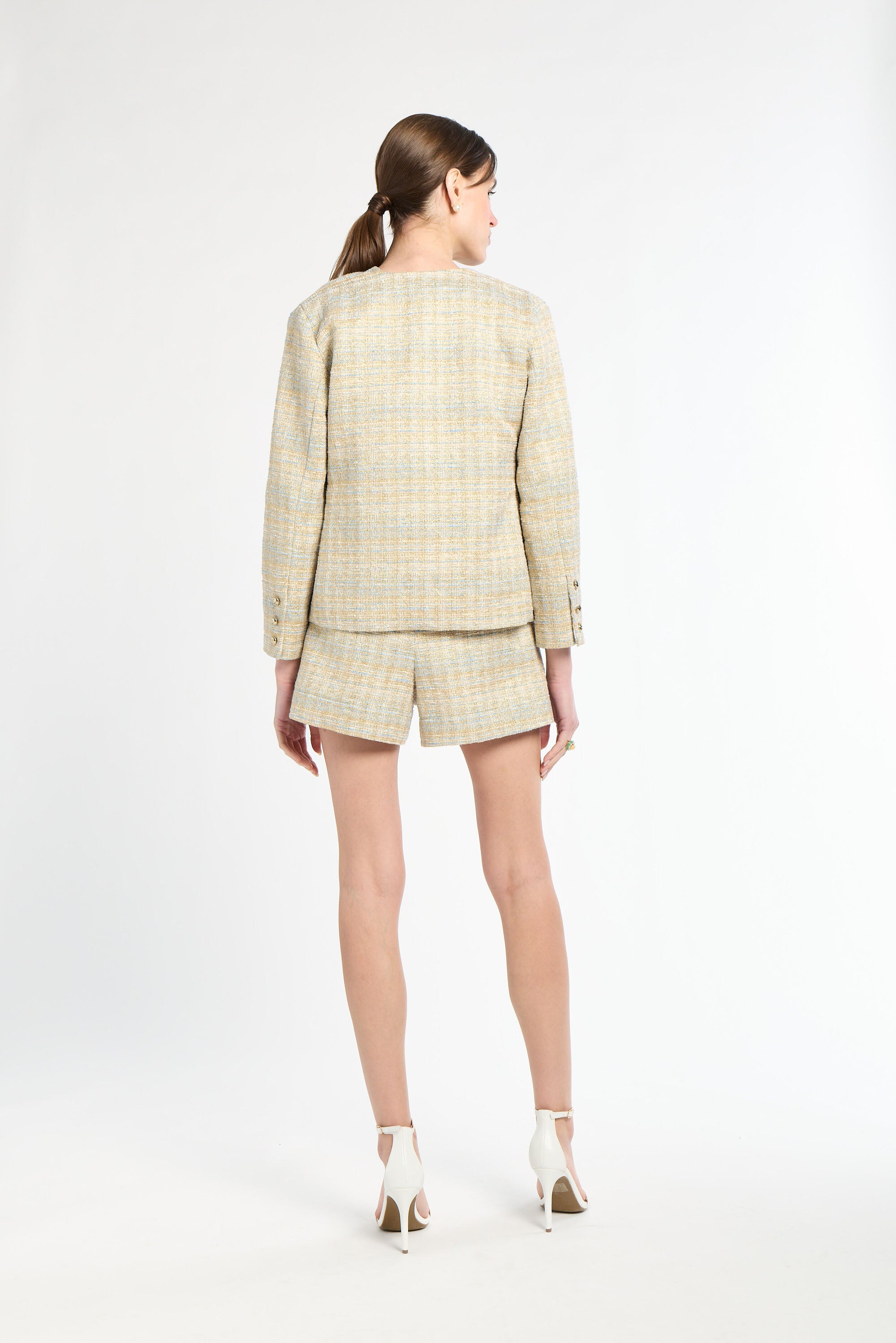 The back of a woman wearing a golden and light blue blazer, shorts, and white heels on a white background