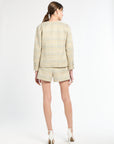 The back of a woman wearing a golden and light blue blazer, shorts, and white heels on a white background