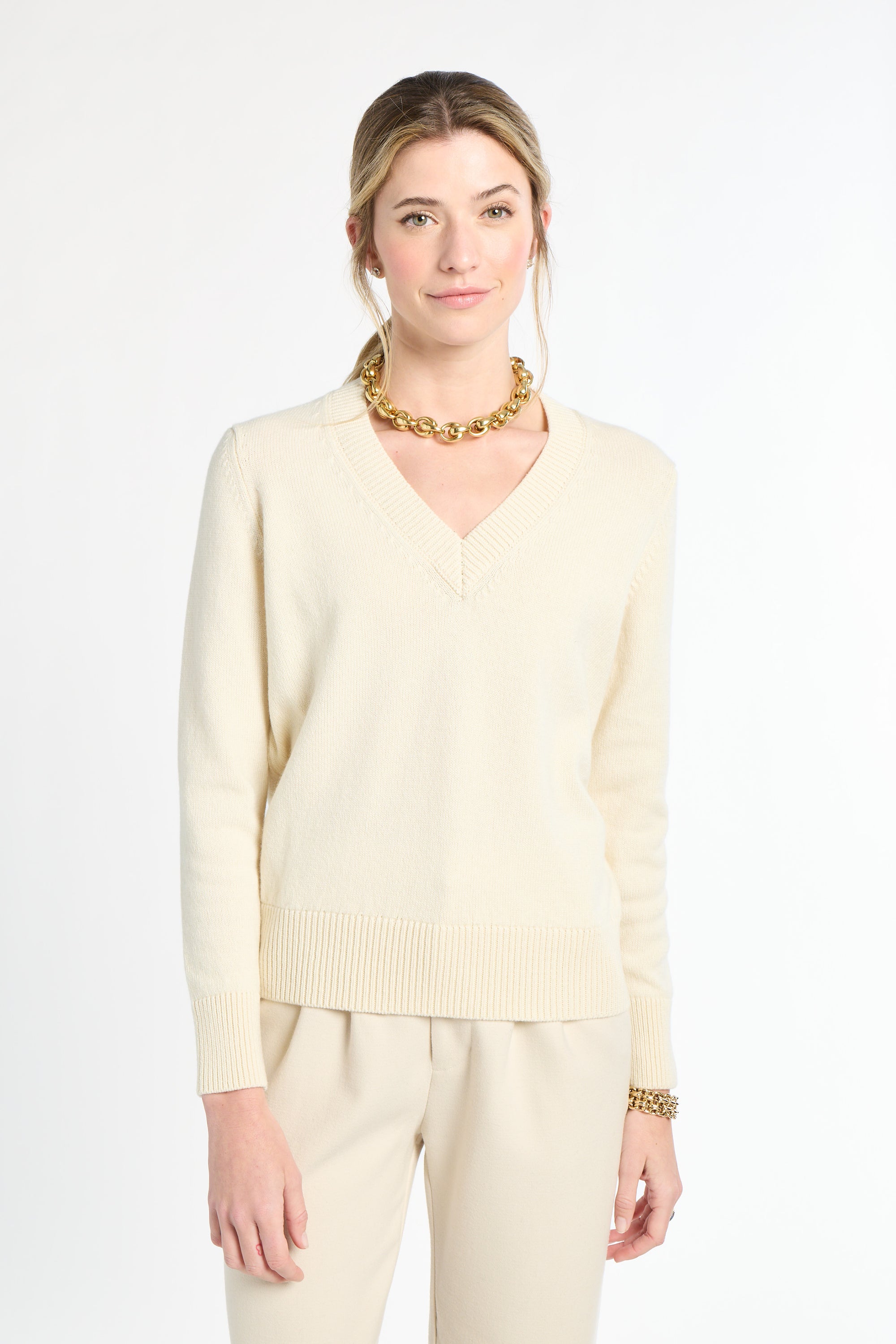 Woman wearing a light-colored sweater and pants with a gold necklace on a white background